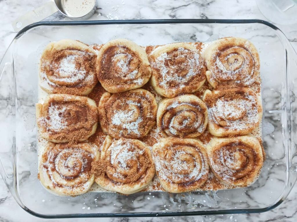 Rolled raw cinnamon rolls in pan with sprinkled cinnamon sugar and warm cream